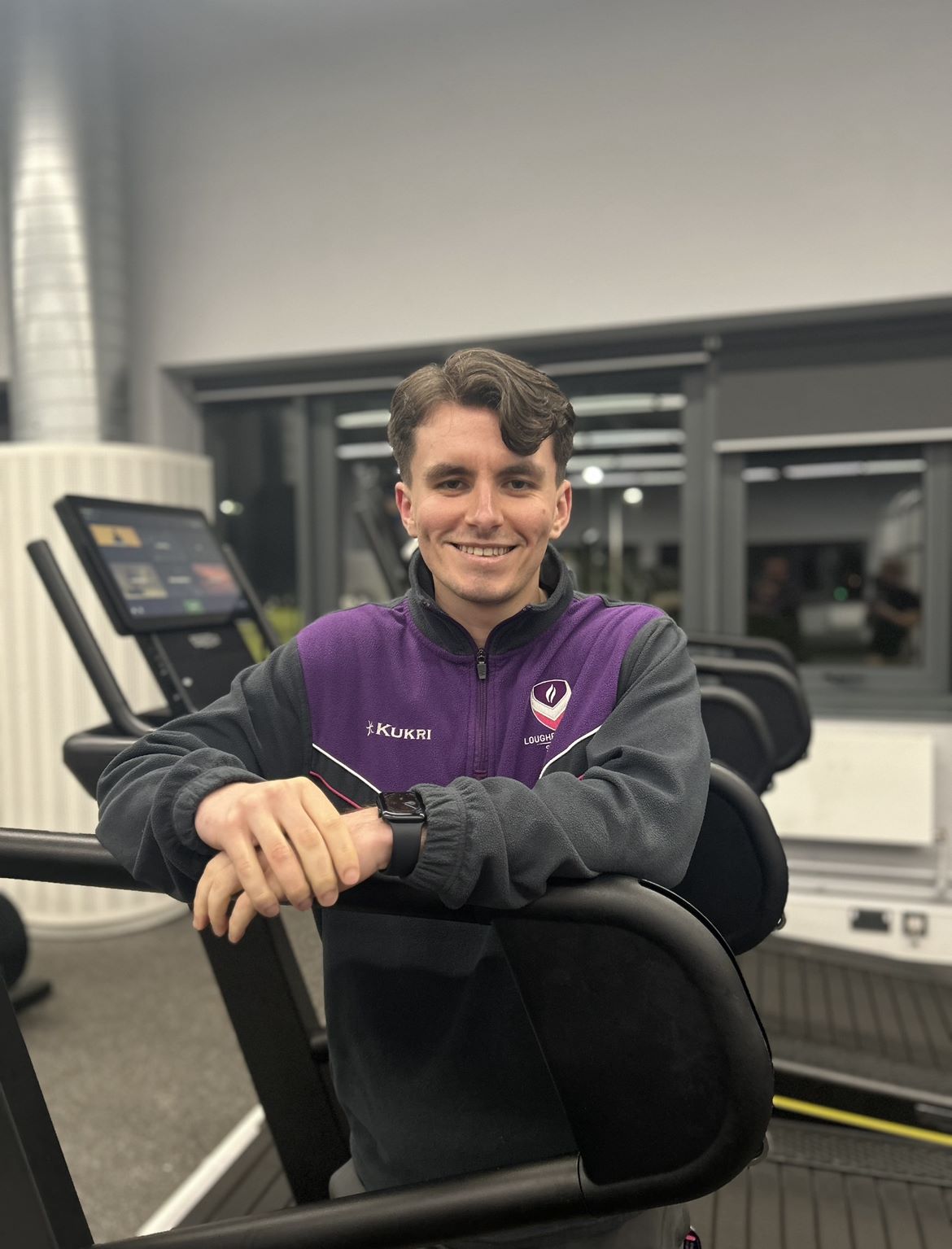 Fitness Coach, Jake, smiling and leaning on a treadmill in Holywell Fitness Centre
